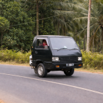 Mobil mini pickup hitam lucu melaju di jalan pedesaan dengan latar pohon kelapa